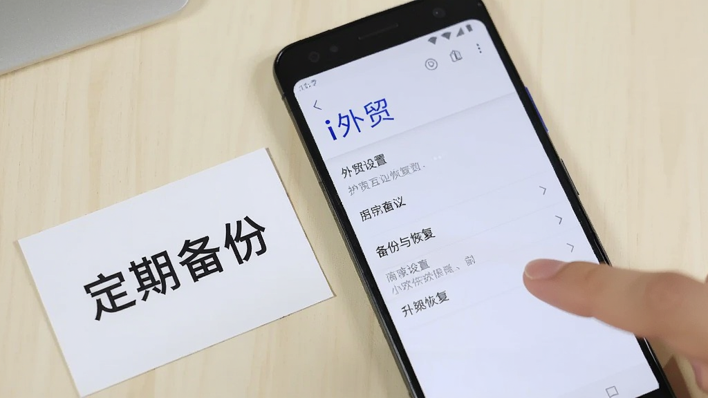 一部安卓手机屏幕显示“i外贸”设置页面中的备份与恢复选项,旁边放置一张写有“定期备份”字样的提示卡片和一只手指向屏幕,背景为浅色桌面,风格为简洁明快的数码产品使用场景图,构图聚焦手机屏幕,光线均匀,画质清晰,画面干净无冗余元素